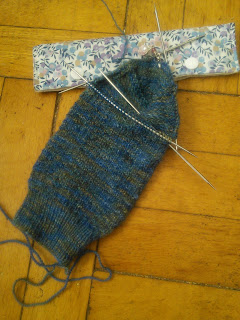 A sock on double-pointed needles, with the heel turn completed. The sock leg is textured, and knit with fingering weight yarn.  The yarn is blue and grey with flecks of brown and yellow. There's a stitch marker with a shell charm clipped into the top of the heel. The heel of the sock is resting on a fabric double-pointed needle case. 