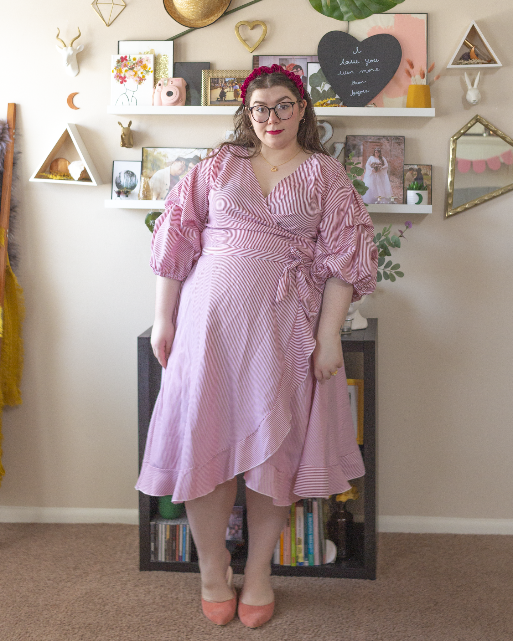 An outfit consisting of a burgundy velvet headband, a pink and white thin vertically striped 3/4 sleeve puffy sleeve wrap midi dress and pastel pink pointed toe heeled clogs.