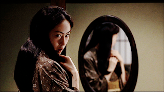 Uma moça japonesa em frente a um espelho, vira a parte superior do corpo para olhar ameaçadoramente para trás (olhando onde está a câmera), segurando com as duas mãoes uma mecha de seu longo e liso cabelo negro.