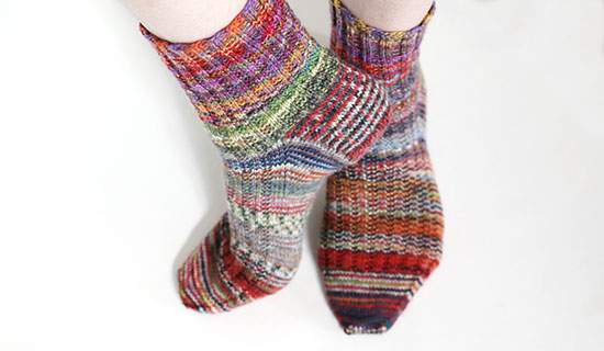 Casually posed feet wearing striped multi-colored hand knit wool socks on a white background.