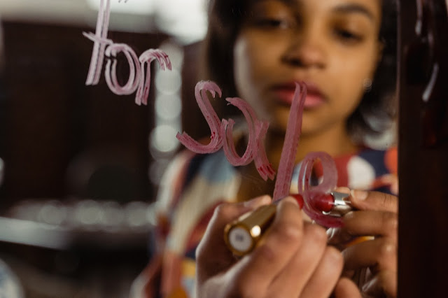 A woman writing "FOR SALE" on a mirror using a lipstick.