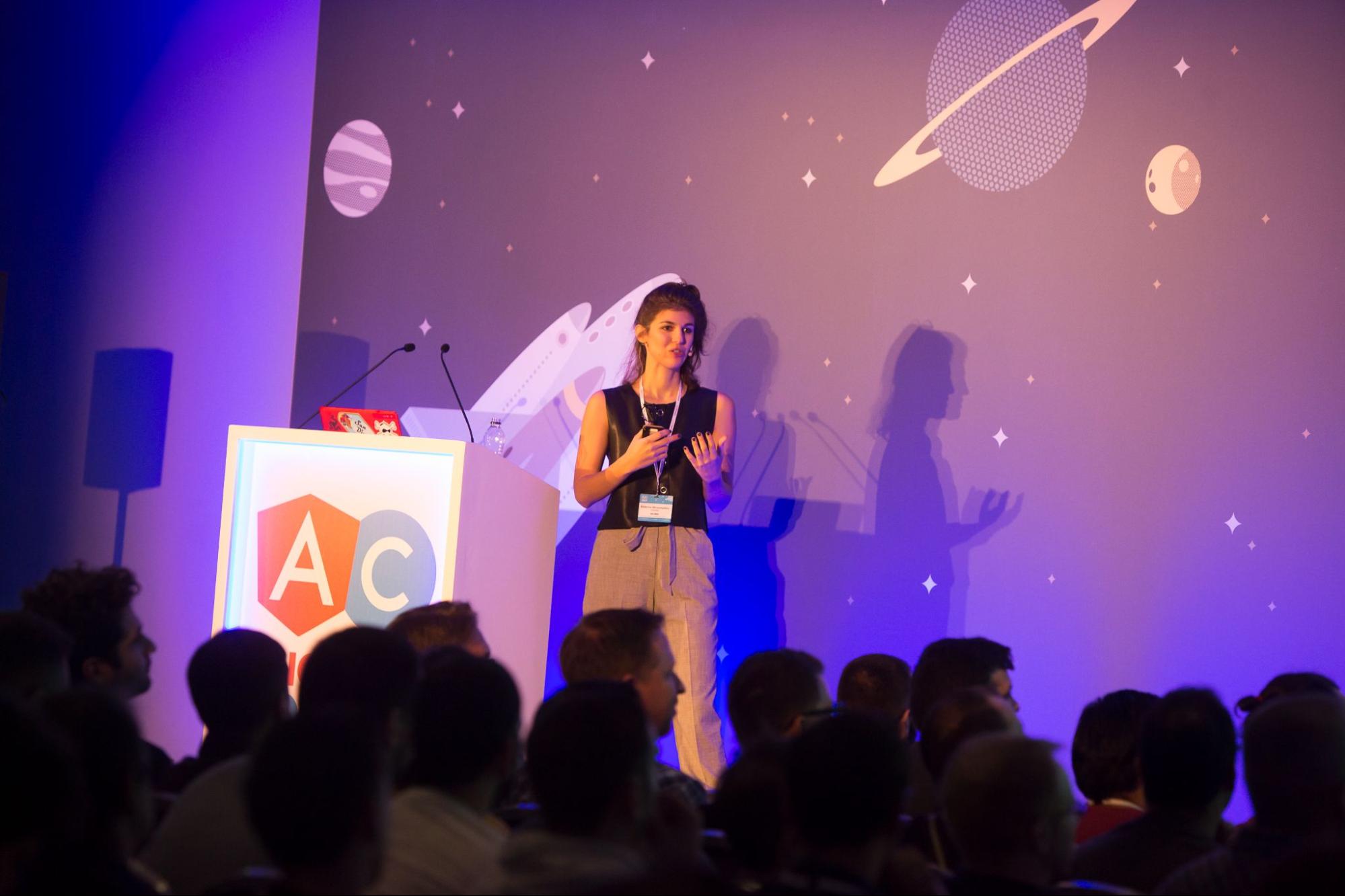 Image shows Katerina Skroumpelou presenting onstage at a conference. Behind her is a podium and a wall covered in a planetary theme