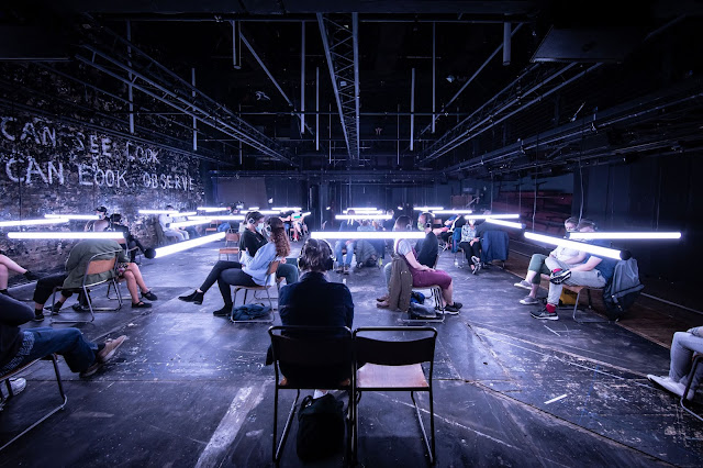 A photo of stripped down theatre space. Audience members sit in spaced-out pairs of chairs, each wearing a large set of headphones. Light fixtures shaped like thin, long tubes have been lowered to face level and hang in the spaces between audience chairs. On the bare brick wall, the words "CAN SEE LOOK" and underneath, "CAN LOOK. OBSERVE." are written in white paint.