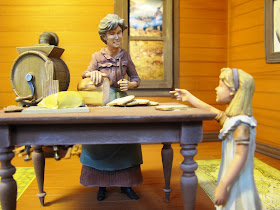 Model of a 19th-century woman and child inside a house. The woman is cutting a loaf of bread, and the child is pointing at a pat of butter.