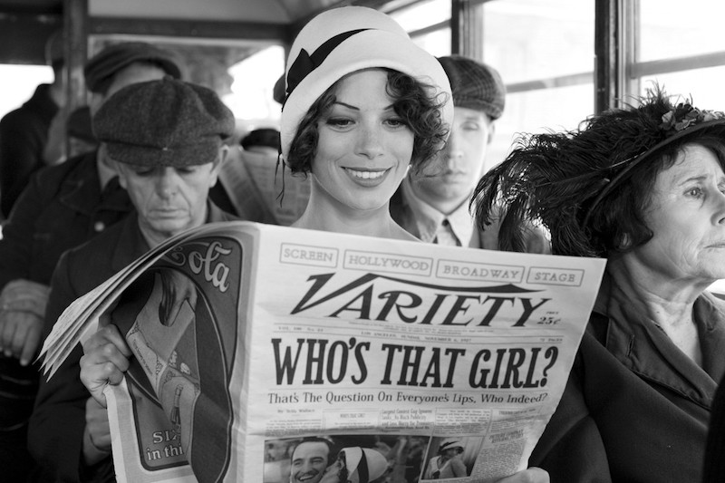 Bérénice Bejo as Peppy Miller, a pretty young woman with short curled hair under a hat, smiling as she sits on a trolley reading 'Variety' with the headline 'Who's That Girl?'
