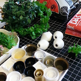 A tray of various bottle caps, a tray of various bit of plastic plant, two wreaths of leaves, a bag of modelling scenic material and a block af DAS air-drying clay arranged on a cutting mat.
