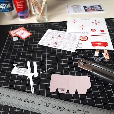 Half-cut out 1/12 scale miniature paper kit on a cutting board, with ruler, cutting knife, glue and instructions.
