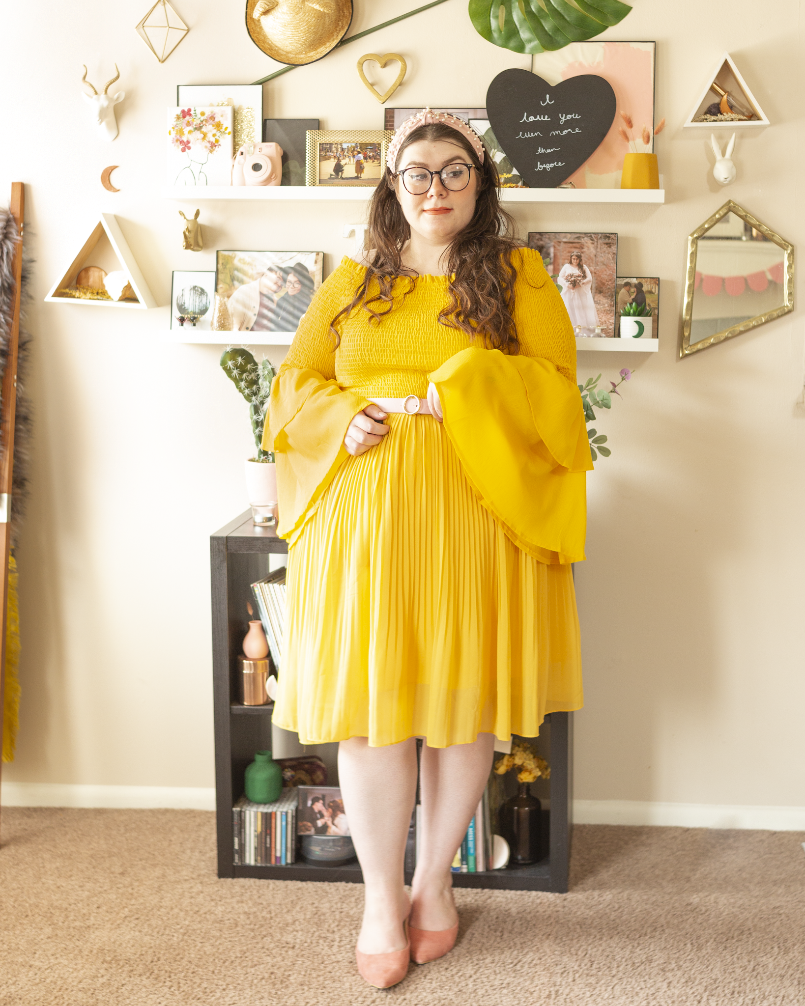 An outfit consisting of a pastel pink pearl encrusted headband, a yellow shirred off the shoulder blouse tucked into a yellow pleated midi skirt, belted with a pastel pink belt and pastel pink heeled clogs.