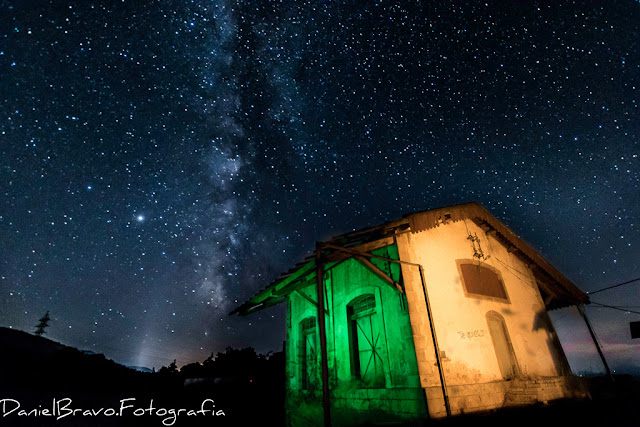 Fotografía nocturna de la estación abandonada de Aldeanueva del Camino en Cáceres donde se ve un cielo estrellado con la Vía Láctea y la estación pintada con luz de dos colores