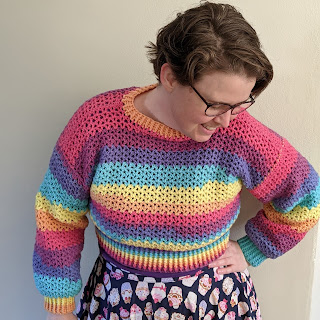 Sarah is standing against a wall facing the camera, she is wearing a rainbow crochet cropped jumper and a cupcake skirt. She is smiling and looking down and away from the camera, with one hand on hip and the other at her side.
