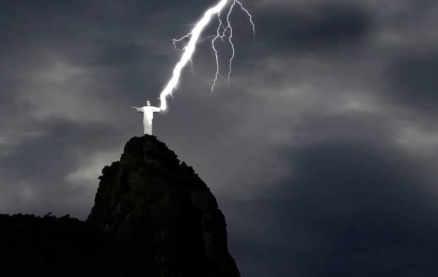 A imagem mostra o cristo redentor sendo atingido por um grande raio vindo dos céus. Foto escura tirada a noite.