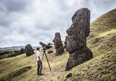 Una persona en la ladera de la montaña, con una cámara fotográfica en un trípode, señala un moai, con otros moais de fondo.