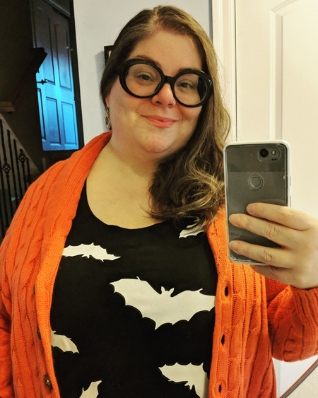 image of me standing in a mirror in my entryway, wearing black-framed glasses, a black t-shirt with white bats on it, and an orange cardigan sweater