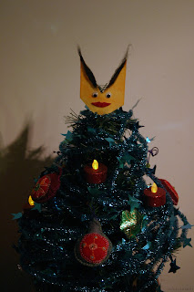 Crude felt face with large dramatic eyebrows, googly eyes, and bright red lips, atop a small yule tree.