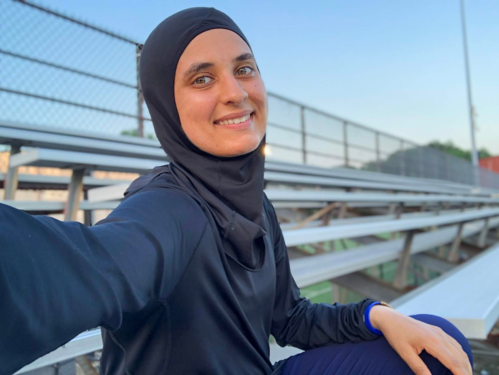 Sahara sitting on a bench at the track, smiling at camera in black nike hijab