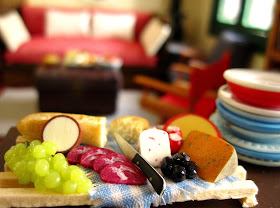 Miniature dolls' house cheese platter on a table next to a pile of plates, In the background is a seating area.