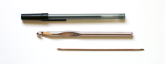 A pen, crochet hook, and double point sock knitting needle aligned for size comparison on a white background.