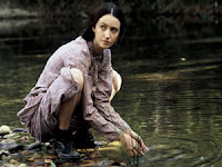 Biela está agachada na beira de um rio, com a saia arregaçada até as coxas, se preparando para levar as mãos à água.