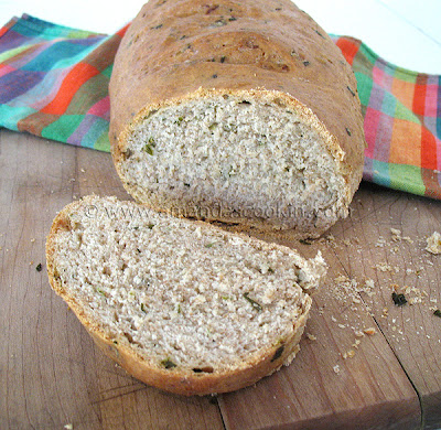 A close up photo of a sliced loaf of scallion chive bread with garlic and rosemary on a wooden cutting board.