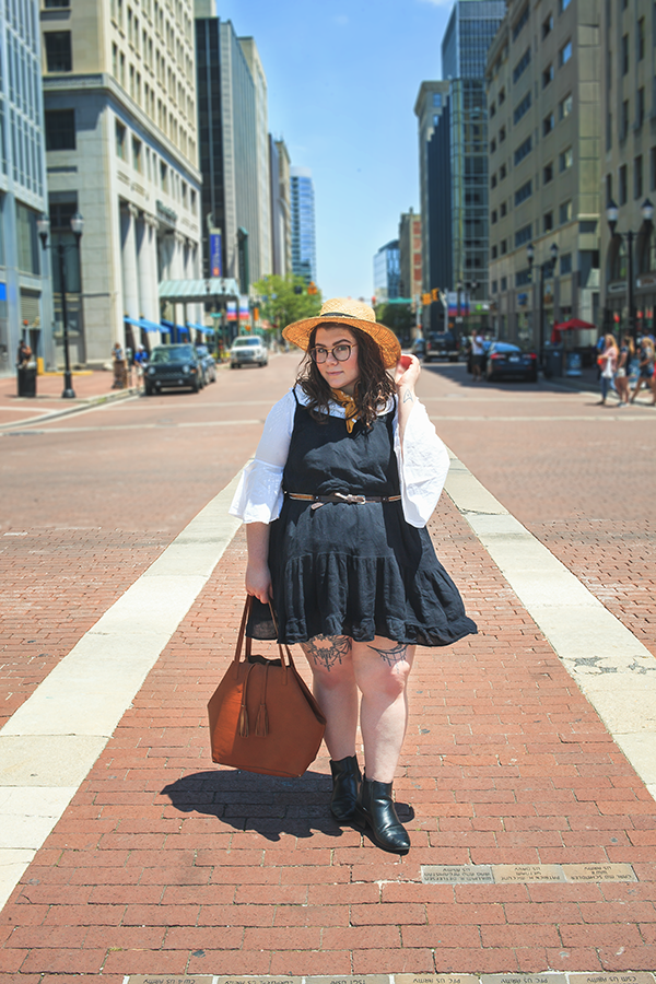 An outfit consisting of a straw boater hat, a white off the shoulder ruffle neck hem blouse, black tiered camisole dress, and black heeled chelsea boots.