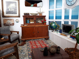1/12 scale modern miniature lounge scene with two mid-century modern armchairs in grey with dark stained arms and legs, an early twentieth century cupboard, a battered sea chest and afghan rug on the floor. On the wall is a framed picutre of a boat and an abstract landscape. On the windowsill is a globe, several piles of books, some shells, a telescope and a flat-screen TV.
