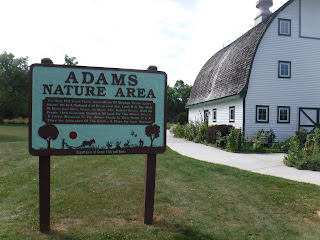 the large white building housing the nature center sits in the background, while a sign in the foreground welcomes visitors to the Adams Nature Area