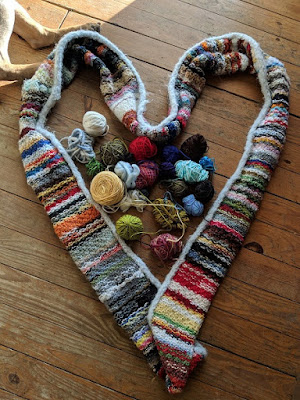 An infinity scarf made from scrap yarn pictured in the shape of heart, with small remnants of yarn in the middle and two dog legs in the upper corner.