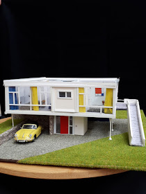 External view of a 1/48th scale model of a mid-century modern house. with a yellow car parked underneath, and a ramp up the right-hand side. The model is on a turntable, with a black cloth behind.