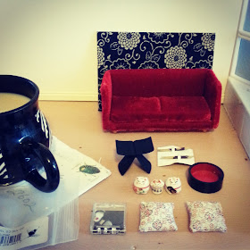 A mug of tea on a used envelope in a one-twelfth scale miniature room. Next to the mug is a card with a piece of blue and white japanese fabric on its front, a deep red miniature velvet sofa, a one-twelfth scale Yanagi butteryfly stool, three one-twelfth scale lucky cats, two one-twelfth scale cherry-blossom cushions, a pair of one-twelfth scale slippers and two pairs of one-twelfth scale chop sticks.