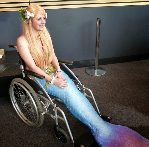 Photo of young woman with long blonde hair and a mermaid costume on her lower body - she is seated in a wheelchair
