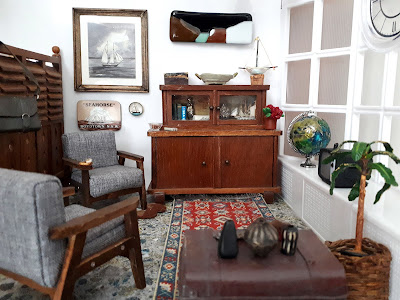 1/12 scale modern miniature lounge scene with two mid-century modern armchairs in grey with dark stained arms and legs, an early twentieth century cupboard, a battered sea chest and afghan rug on the floor. On the wall is a framed picutre of a boat and an abstract landscape. On the windowsill is a globe. 