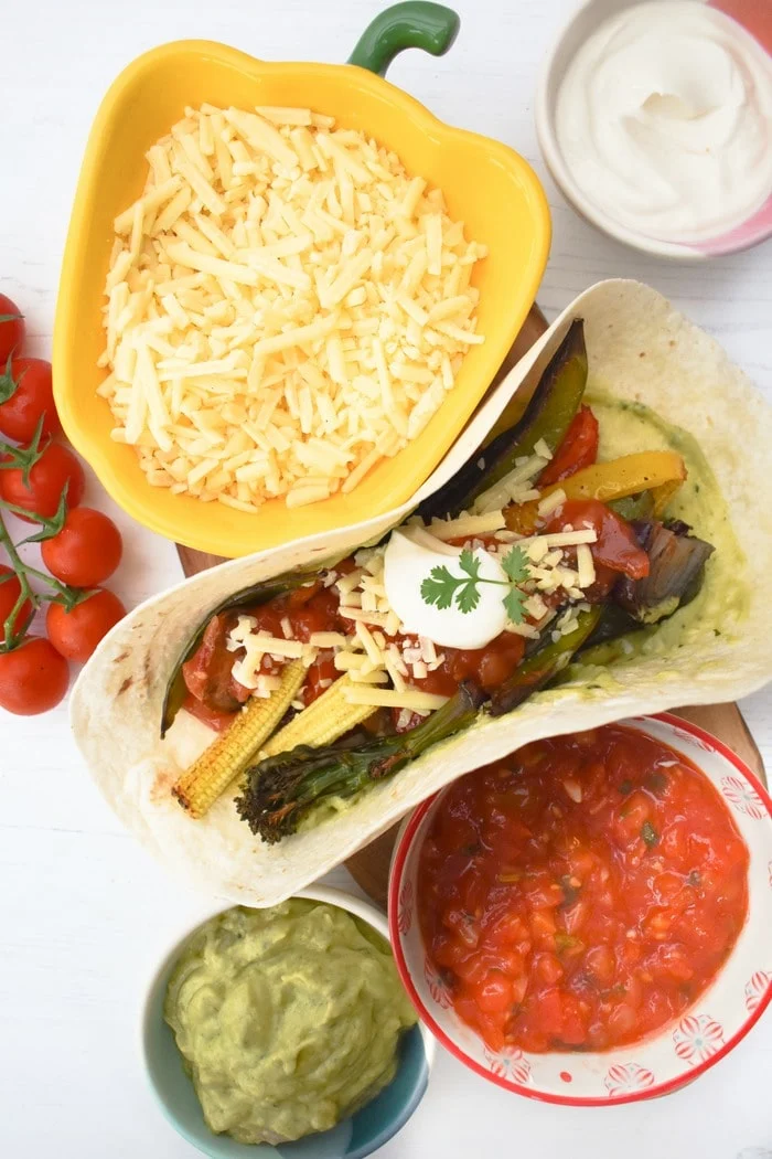 Vegetable fajita surrounded by bowls of salsa, guacamole, grated cheese and cherry tomatoes