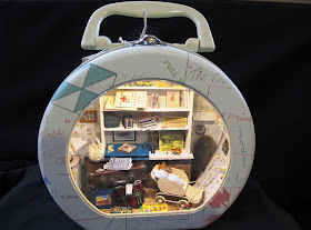 Small round metal suitcase with a clear front, containing a one-twelfth scale scene containing 1940s items.