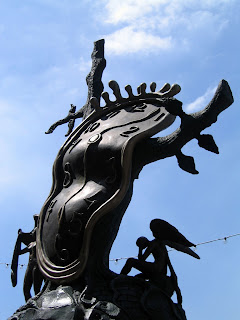 A sculpture of a warped clock with an angel in the background with its head in its hand.