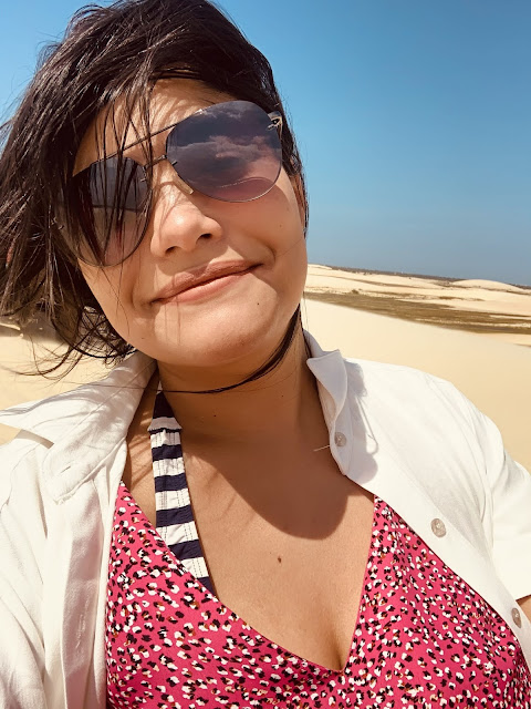 mulher morena, jovem, cabelo preto, oculos escuros, blusa rosa e blaser branco em uma duna de areia