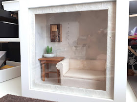 White box frame containing a 1/12 scale modern miniature cream sofa, with a wooden table next to it. On the table is a potted cactus, and above it is a book-themed piece of assemblage art.
