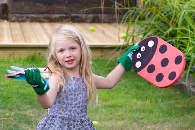 A 4 year old girl in a dress with green gardening gloves, gardening tools and a ladybird kneeling map