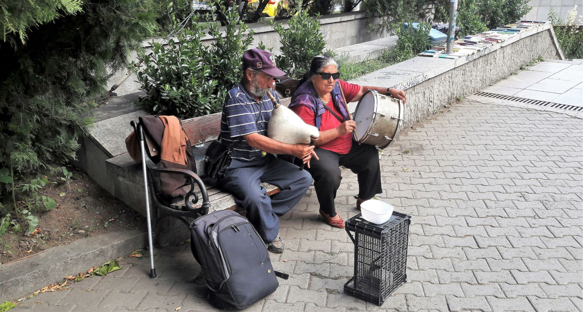 An man playing the pipe and a woman playing the drum while seating on a bench