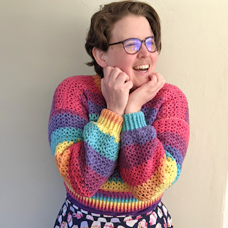 Sarah is wearing a rainbow crochet crop jumper with v-stitch. She is looking off to the side and laughing, with her hands under her chin.