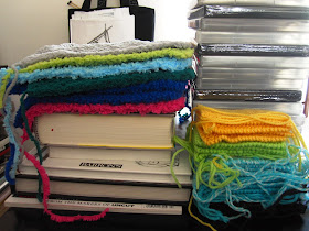 Piles of books, DVDs and rainbow-coloured stacks of knitted rectangles with trailing yarn ends
