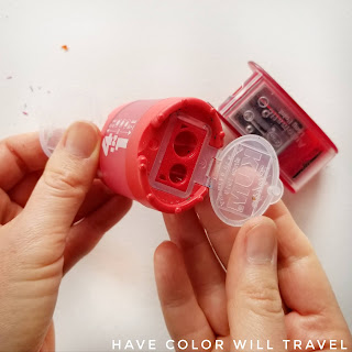 The red KUM 4-in-1 pencil sharpener held in a woman's hand up close, and the lid of the blade cover is open.