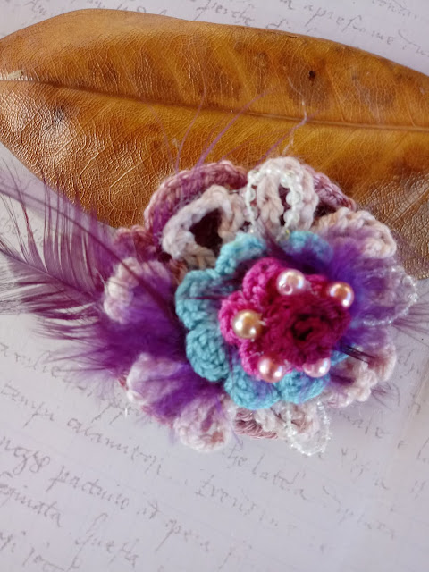 Flor tejida a ganchillo de varios colores y decorada con plumas violeta y perlas de color rosa
