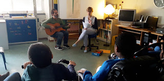 A professora conta sentada a história acompanhada do professor que toca uma guitarra e os alunos escutam com atenção.