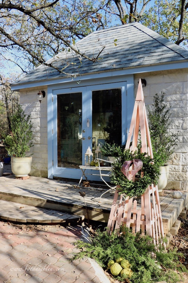 A peach colored trellis decorated with a boxwood wreath and foraged cedar branches and horse apples sits at the edge of the garden shed's deck