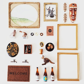 Flat lay of one-twelfth miniature items in black, brown and gold including picture frames, wall art, a sewing kit, wooden fruit and bottles of wine