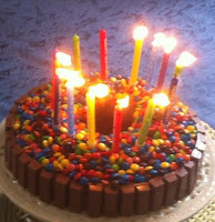 Home-cooked birthday cake: chocolate sponge ring cake with kit-kat sides to contain the candy buttons covering the top. Most of the birthday candles standing in a ring are topped with flame but a few have been blown out already. May wishes come true!