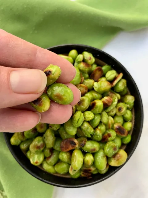 Bowl of finished edamame with a hand holding three soybeans.
