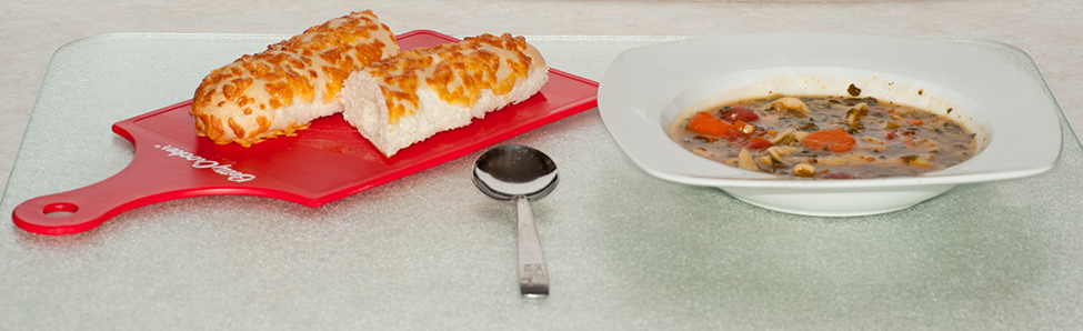 A bowl of homemade veggie soup served with a long narrow cheese bun.