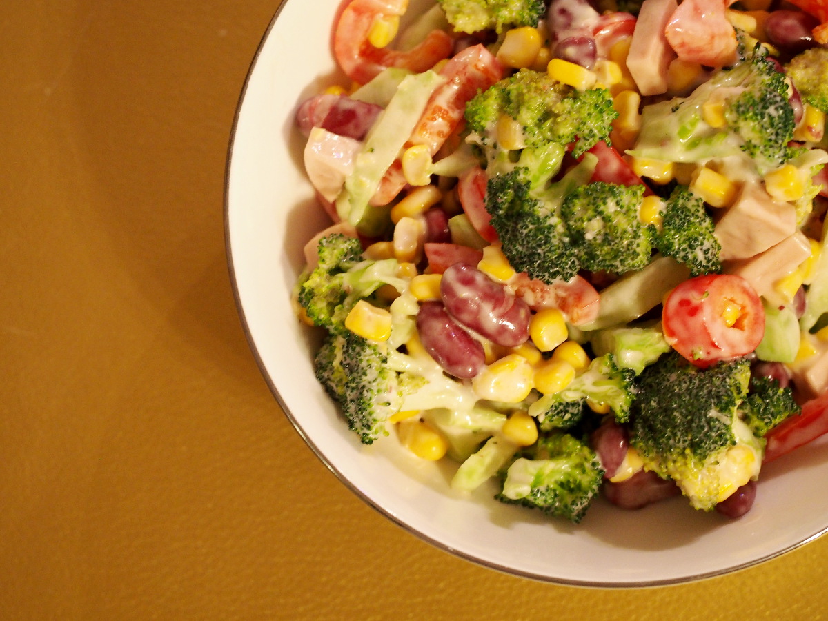 Eine Schüssel, gefüllt mit dem Brokkolisalat. Man sieht die einzelnen Bestandteile: den grünen Brokkoli, den gelben Mais, die roten Kidneybohnen, die rote Paprika und Stücke der veganen "Fleischwurst".