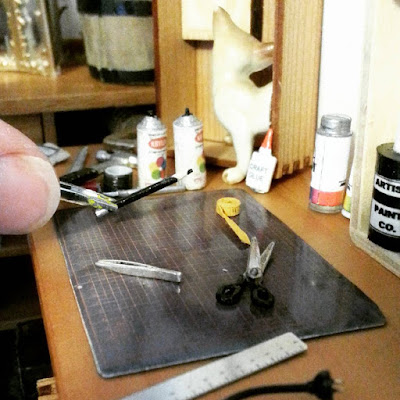 1/12 scale studio scene with a workbench containing a cutting mat, glue, scissor, ruler, paint and glue. At the back is a workbench full of supplies. A full-sized thumb is in the bottom left corner, holding a pair of 1/12 tweezers, holding a 1/12 pencil.